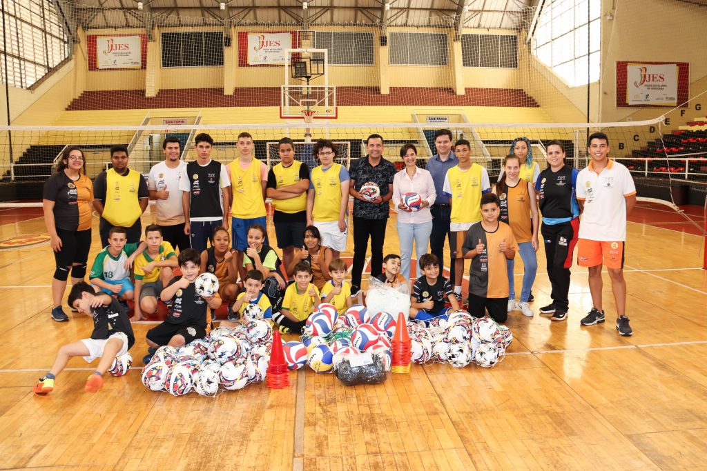 Prefeita Jaqueline com o secretário de esportes Simei Lamarca e as crianças participantes do projeto Jovem Talento, em uma quadra, com bolas de futebol e uma rede de vôlei ao fundo