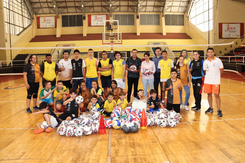 Prefeita Jaqueline com o secretário de esportes Simei Lamarca e as crianças participantes do projeto Jovem Talento, em uma quadra, com bolas de futebol e uma rede de vôlei ao fundo