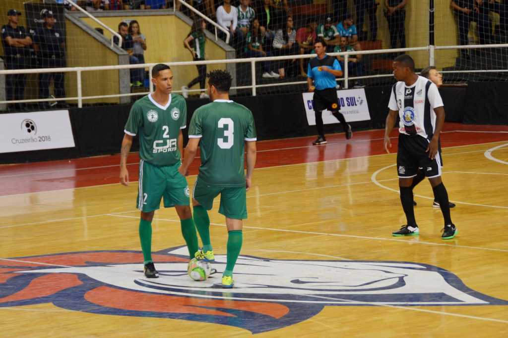 jogadores de futsal no meio da quadra.
ambos de uniforme verde.
em deles com a bola no pé, seguido pelo adversário que usa uniforme branco e preto.