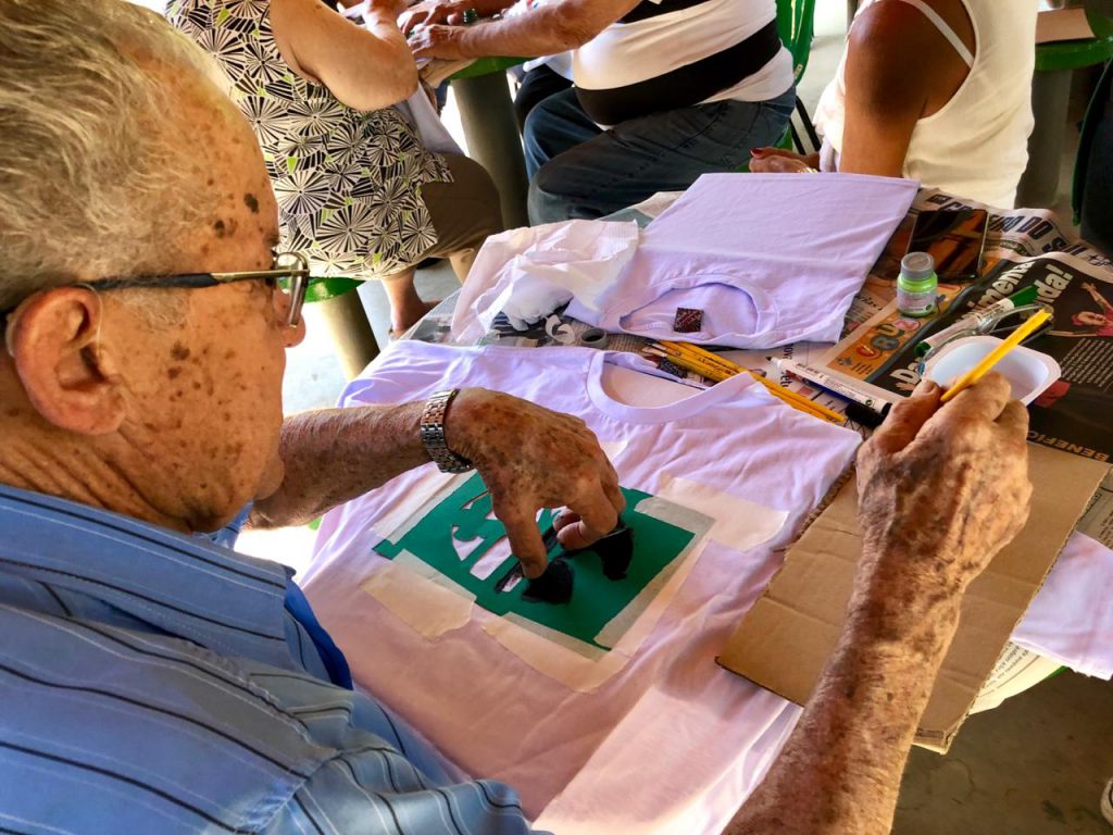 Idoso sentado fazendo pintura em camiseta.
está utilizando a cor preta.
